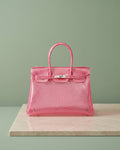 Croco Glacé Fuchsia, sac à coque brillante effet croco en rose framboise, posé sur du marbre devant un mur sauge